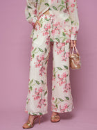 A person poses against a pink background wearing Social Queen’s Floral Embroidered Textured Wide-Leg Pants with a matching top, styled with a small bag and gold high-heeled sandals.