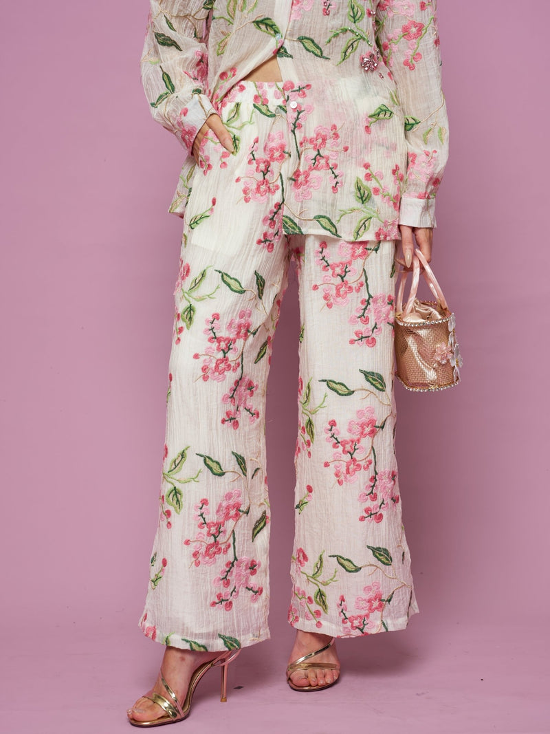 A person poses against a pink background wearing Social Queen’s Floral Embroidered Textured Wide-Leg Pants with a matching top, styled with a small bag and gold high-heeled sandals.