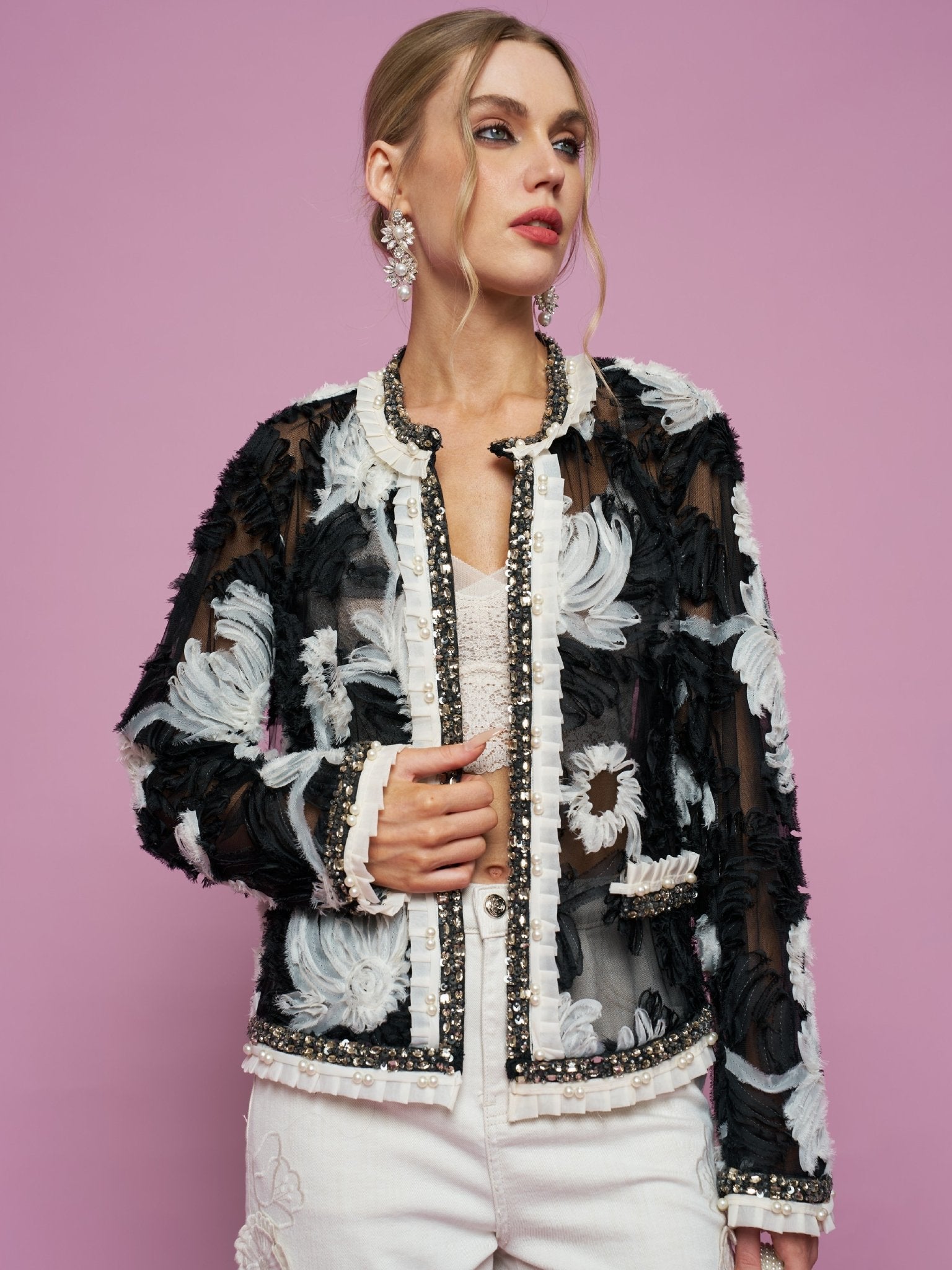Against a pink background, a woman wears the Social Queen Floral Embroidered Tulle Beaded Jacket over a white top and pants, styled with statement earrings.
