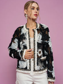 Against a pink background, a woman wears the Social Queen Floral Embroidered Tulle Beaded Jacket over a white top and pants, styled with statement earrings.