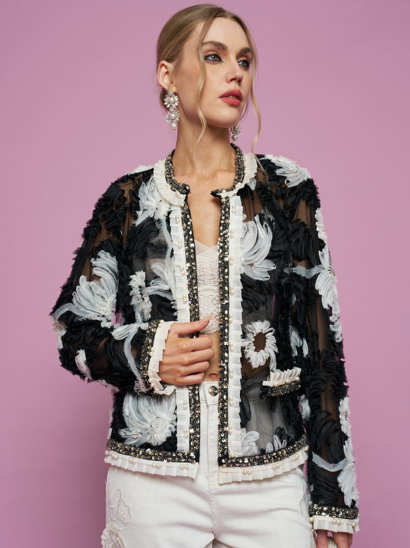 Against a pink background, a woman wears the Social Queen Floral Embroidered Tulle Beaded Jacket over a white top and pants, styled with statement earrings.