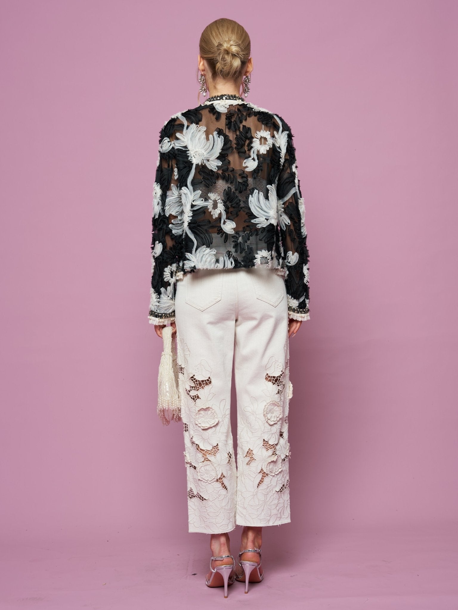 A woman stands facing away against a pink background, wearing the Social Queen Floral Embroidered Tulle Beaded Jacket with white embroidered pants, pink heels, and holding a fringed purse.