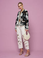 A woman stands against a pink background wearing Social Queen’s Floral Embroidered Tulle Beaded Jacket, white embroidered pants, silver heels, and holding a beaded white handbag.