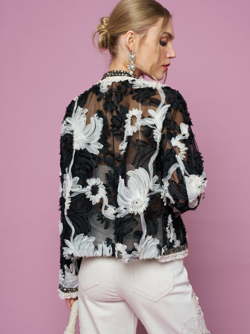 A woman stands against a pink background, facing away and wearing the Social Queen Floral Embroidered Tulle Beaded Jacket paired with white pants, highlighting the jacket's sheer mesh and intricate 3D floral embroidery.