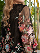 A person with long, wavy blonde hair is wearing the Social Queen Floral Embroidery 100% Mulberry Silk Tulle Blouse, a sheer black top adorned with colorful flowers, standing before green plants.