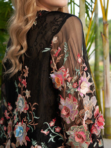 A person with long, wavy blonde hair is wearing the Social Queen Floral Embroidery 100% Mulberry Silk Tulle Blouse, a sheer black top adorned with colorful flowers, standing before green plants.