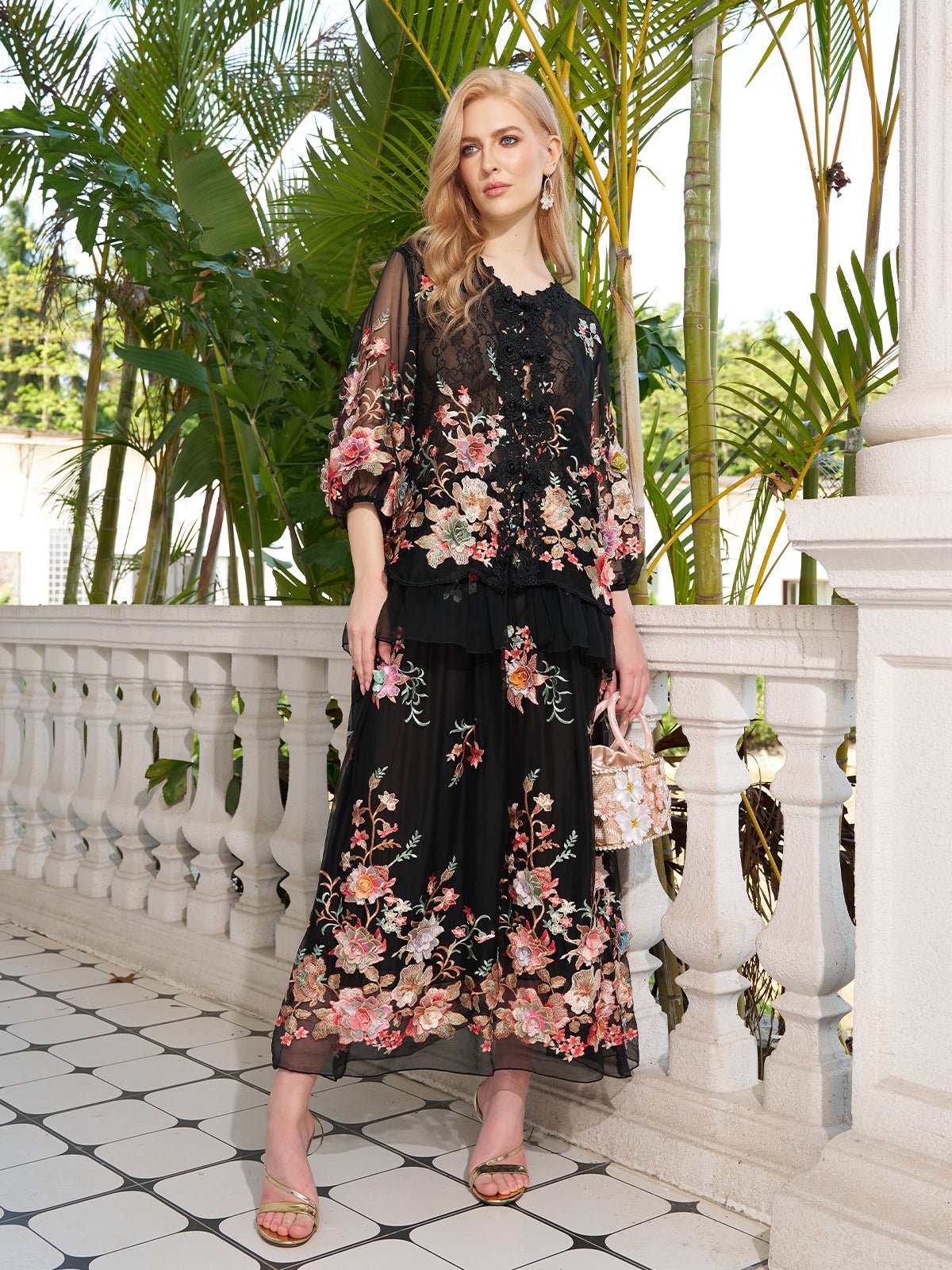 A woman stands on a tiled balcony with greenery, wearing the Social Queen Floral Embroidery 100% Mulberry Silk Tulle Blouse paired with a long black floral dress, sandals, and holding a small handbag.
