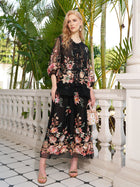 A woman stands on a tiled balcony with greenery, wearing the Social Queen Floral Embroidery 100% Mulberry Silk Tulle Blouse paired with a long black floral dress, sandals, and holding a small handbag.