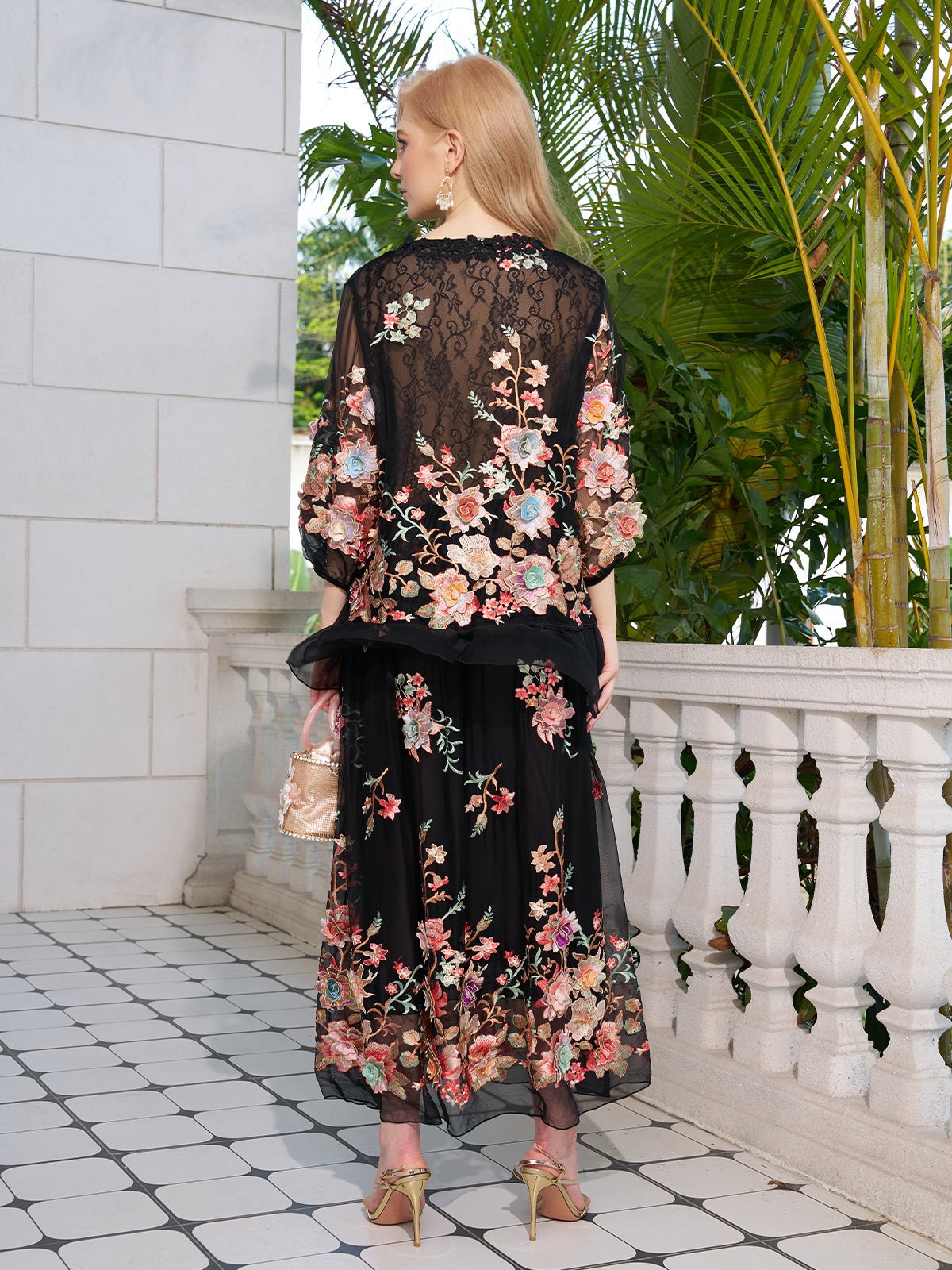 A woman stands on a tiled terrace with her back to the camera, wearing Social Queen's Floral Embroidery 100% Mulberry Silk Tulle Blouse and holding a small handbag. Lush green plants fill the background.