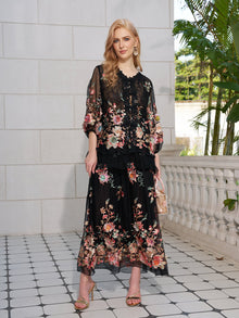 A woman on a tiled terrace wears the Social Queen Floral Embroidery 100% Mulberry Silk Tulle Skirt, a sheer black top, gold sandals, and holds a small handbag. White stone railing and greenery are visible in the background.