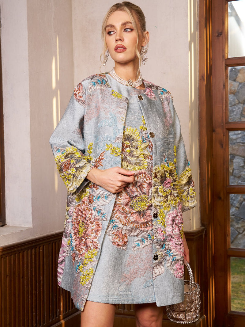 A woman with blonde hair tied back wears the Social Queen Floral Jacquard Beaded Jacket indoors, paired with a matching dress, statement earrings, and a beaded handbag.