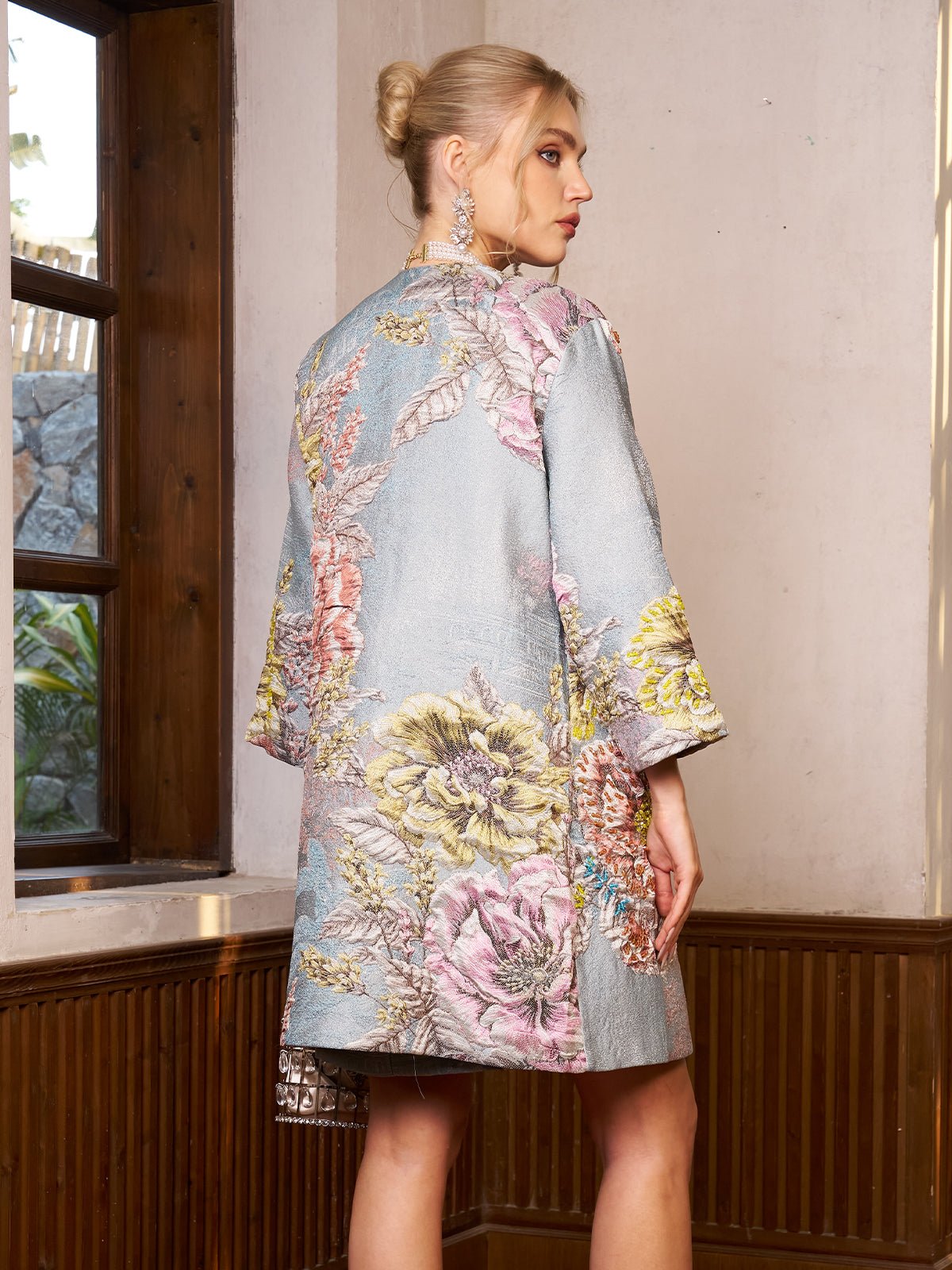 A woman with blonde hair in a bun wears the Social Queen Floral Jacquard Beaded Jacket and large earrings, standing indoors by a wooden window, facing slightly away from the camera.