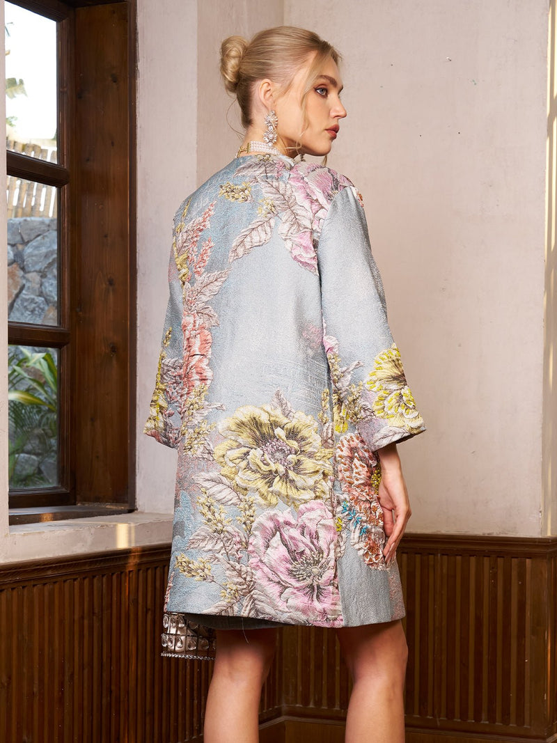 Indoors by a window, a person with hair in a bun and large earrings wears the Social Queen Floral Jacquard Beaded Jacket in light blue.