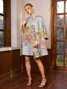 A woman stands indoors in the Social Queen Floral Jacquard Beaded Sleeveless Dress with a matching cape, holding a metallic handbag and wearing heeled sandals—an elegant wedding guest look.