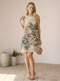 A woman stands indoors in the Social Queen Floral Jacquard Dress, a sleeveless sheath with bold floral patterns, paired with a small woven bag—an elegant wedding guest look accented by vases and flowers in the background.
