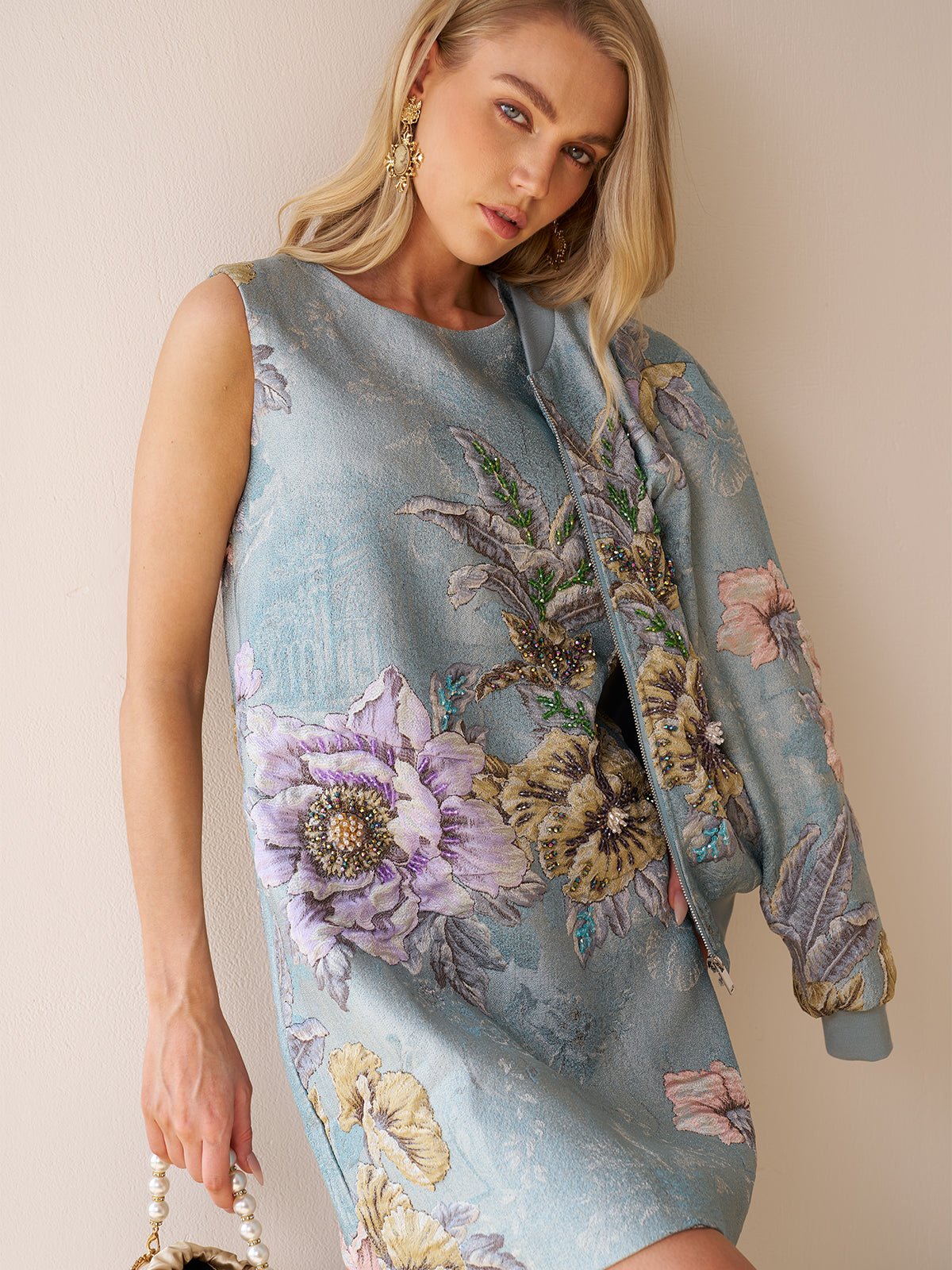 A woman in the Social Queen Floral Jacquard Dress with a matching jacket draped over her shoulder and a beaded bag poses against a neutral wall—a stunning wedding guest look.