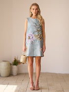 A woman stands indoors wearing the Social Queen Floral Jacquard Dress—a sleeveless sheath with light blue floral embroidery—holding a beaded handbag amid neutral décor and potted plants, making it a lovely choice for a wedding guest.