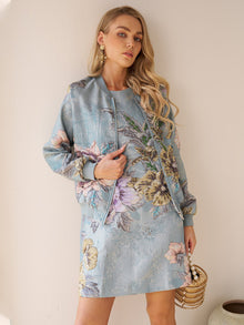 A woman stands against a beige wall in the Social Queen Floral Jacquard Dress, paired with a light blue floral-patterned jacket and a round beaded handbag—an elegant choice for wedding guests.