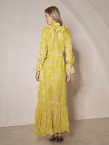 A woman stands indoors with her back to the camera, wearing the Social Queen Floral Jacquard Velvet Ruffles Dress—a long wedding guest dress with ruffled details, long sleeves, and a wrap silhouette—paired with high-heeled shoes.