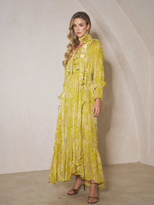 A woman stands indoors wearing the Social Queen Floral Jacquard Velvet Ruffles Dress with long sleeves, ruffles, and a tie at the neck—an elegant choice for wedding guests. She has long wavy hair and heeled sandals.