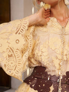 Close-up of a person wearing the Social Queen Floral Lace Embroidered Blouse—a sheer yellow top with 3D floral embroidery and wide sleeves—styled with a brown cut-out leather belt, hand raised near their neck.