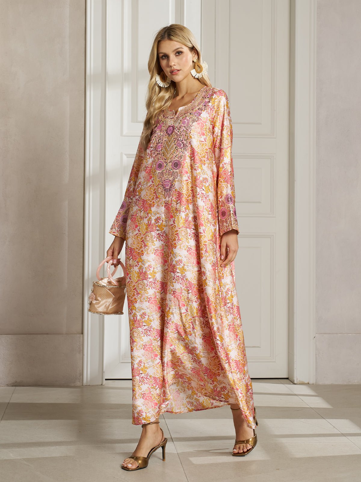 A woman models the Social Queen Floral Print Beaded Dress, a long, loose-fitting robe adorned with a botanical print, while holding a small handbag and standing indoors before white paneled doors.