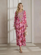 A woman stands indoors in the Social Queen Floral Print Beaded Dress, featuring a long silhouette with a botanical pattern and delicate beadwork, paired with a small handbag and heeled sandals.