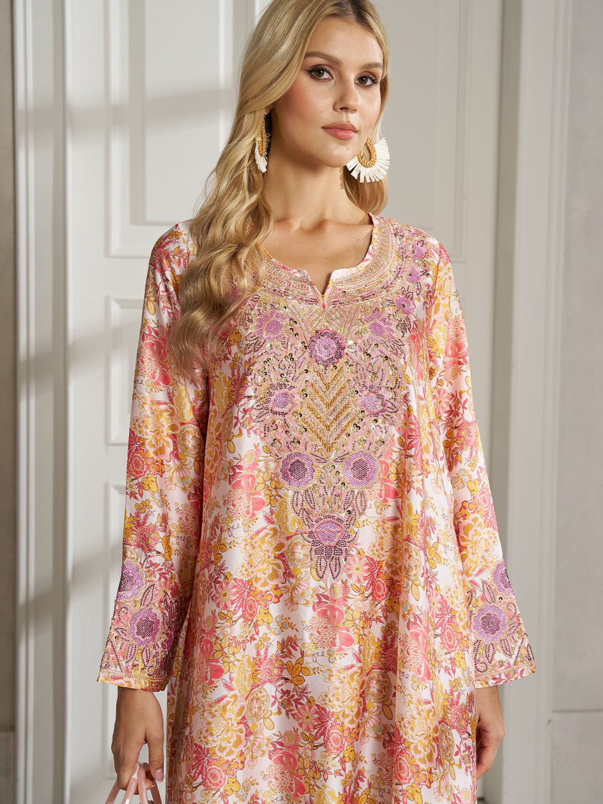 A woman models the Social Queen Floral Print Beaded Dress, featuring long sleeves and a hand-beaded neckline, as she stands indoors against a white paneled wall, gazing slightly to the side.