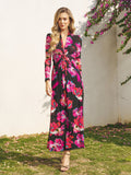A woman stands on grass before a white wall, wearing the Social Queen Floral Print Knot Front Knit Dress—an ankle-length, long-sleeve black dress with bright pink and red florals—styled with beige strappy heels, ideal for wedding guests.