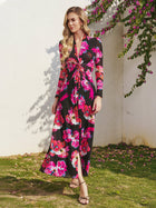 A woman wears the Social Queen Floral Print Knot Front Knit Dress, a long-sleeved black midi with vibrant pink and red florals, while standing on grass near a white wall decorated with hanging pink flowers.