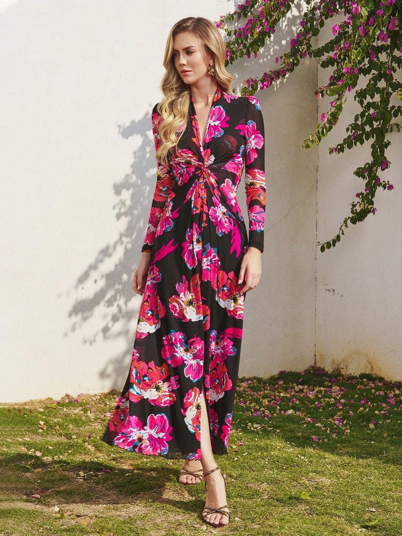 A woman poses outdoors by a white wall and flowering vines, wearing the Social Queen Floral Print Knot Front Knit Dress in black with vibrant pink and red blooms, styled with strappy sandals for a vintage-inspired look.