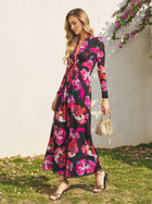A woman stands outdoors on grass in the Social Queen Floral Print Knot Front Knit Dress, featuring long sleeves and pink blooms, paired with a woven handbag and strappy sandals—an elegant wedding guest look.