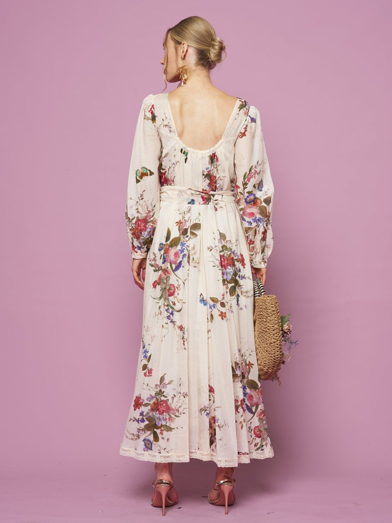 A woman faces away, wearing the Social Queen Floral Print Lace Detail Dress with long sleeves and pink heels, holding a woven bag against a pink background.