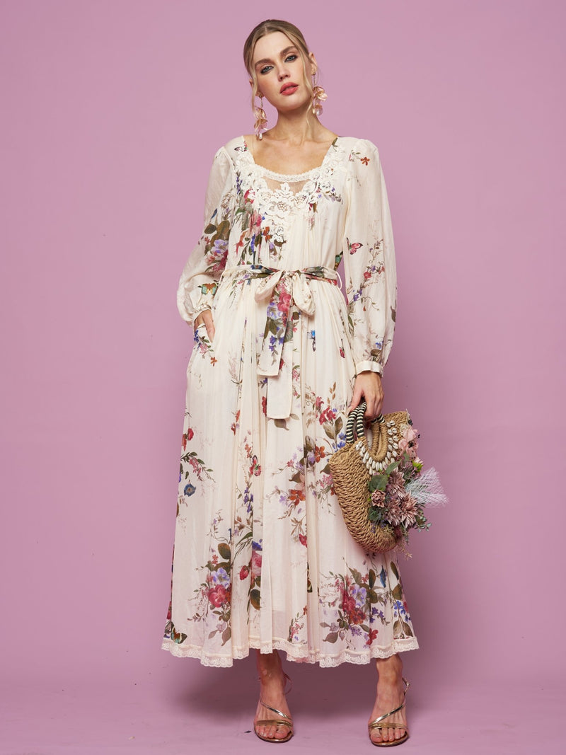 A woman wears the Social Queen Floral Print Lace Detail Dress with long sleeves, standing against a pink background. She holds a flower-adorned straw bag and pairs the look with sandals.