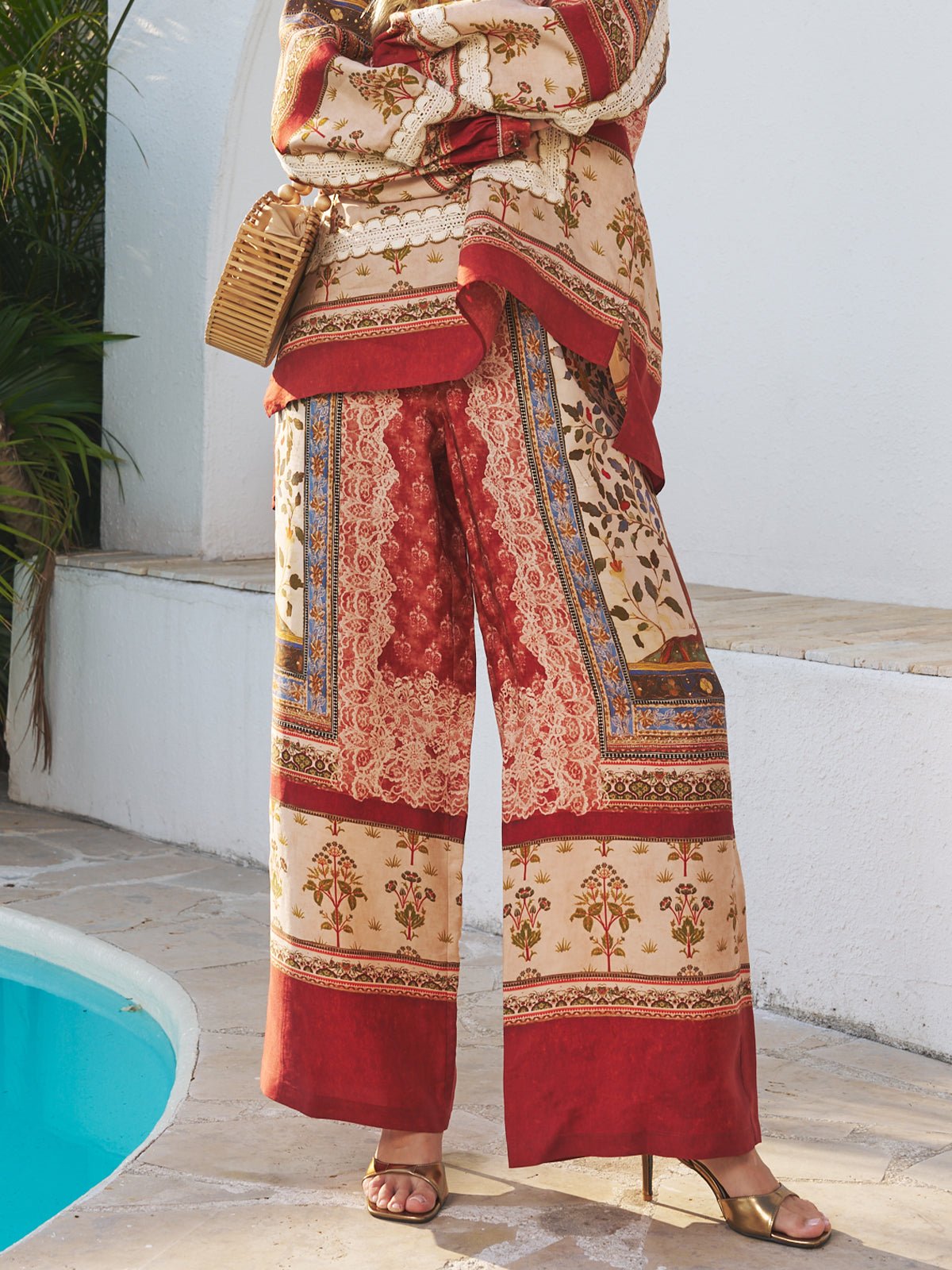 A person stands by a pool in Social Queen Floral Print Wide-Leg Boho Pants and a matching top with botanical and geometric prints, gold heeled sandals, and holding a woven bag.