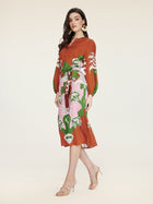 A woman models the Social Queen Floral Printed Silk Dress—rust-colored with long sleeves and green and white florals—paired with nude heeled sandals, exuding vacation elegance against a plain background.