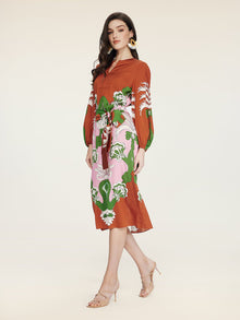 A woman models the Social Queen Floral Printed Silk Dress—rust-colored with long sleeves and green and white florals—paired with nude heeled sandals, exuding vacation elegance against a plain background.