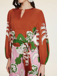 A woman wears the Social Queen Floral Printed Silk Dress, featuring a rust hue, long sleeves, V-neck, and a green and pink tie-waist for effortless vacation elegance.