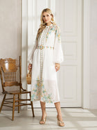 A woman models the Social Queen Floral Printed Stand-Collar Dress, a white, long-sleeved floral dress with a belt, holding a small wicker bag beside a wooden chair—ideal for spring vacations.