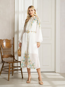 A woman models the Social Queen Floral Printed Stand-Collar Dress, a white, long-sleeved floral dress with a belt, holding a small wicker bag beside a wooden chair—ideal for spring vacations.