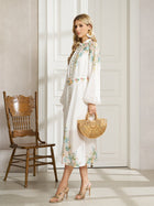 A woman in the Social Queen Floral Printed Stand-Collar Dress pairs it with heeled sandals and holds a bamboo handbag while standing indoors beside a wooden chair.