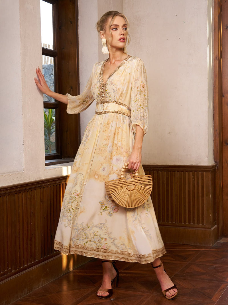 A woman in Social Queen’s Floral Silk V-Neck Beaded Dress stands by a window, accessorized with a bamboo handbag, statement earrings, and black sandals—an elegant choice for any wedding guest.