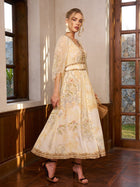 A woman wears the Social Queen Floral Silk V-Neck Beaded Dress, pairing it with high-heeled sandals, statement earrings, and a clutch as she stands by tall windows—perfect for a wedding guest look.