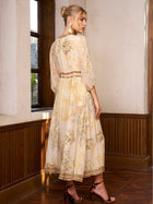 A woman stands indoors by a window in the Social Queen Floral Silk V-Neck Beaded Dress—cream with delicate florals and sheer sleeves—paired with brown heeled sandals, perfect for wearing as a wedding guest.