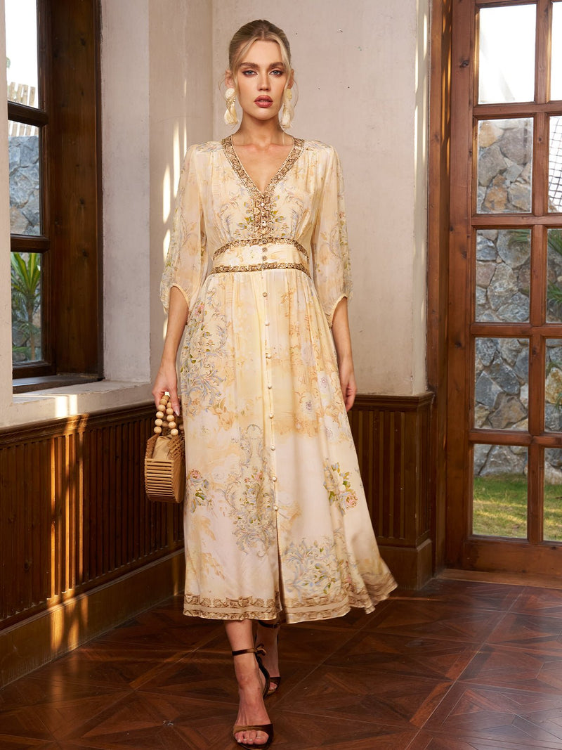 A woman stands indoors by a sunlit window, wearing the Social Queen Floral Silk V-Neck Beaded Dress in light beige with gold accents and subtle floral print, paired with brown heels and holding a woven handbag.