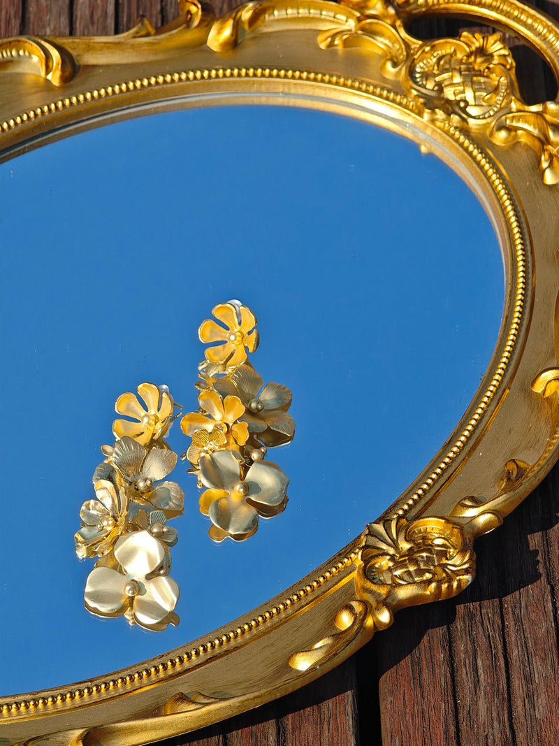 Social Queen’s Flower Tassel Earrings, featuring a layered flower design, are displayed on an ornate gold-framed mirror that reflects the blue sky and wooden surface below.