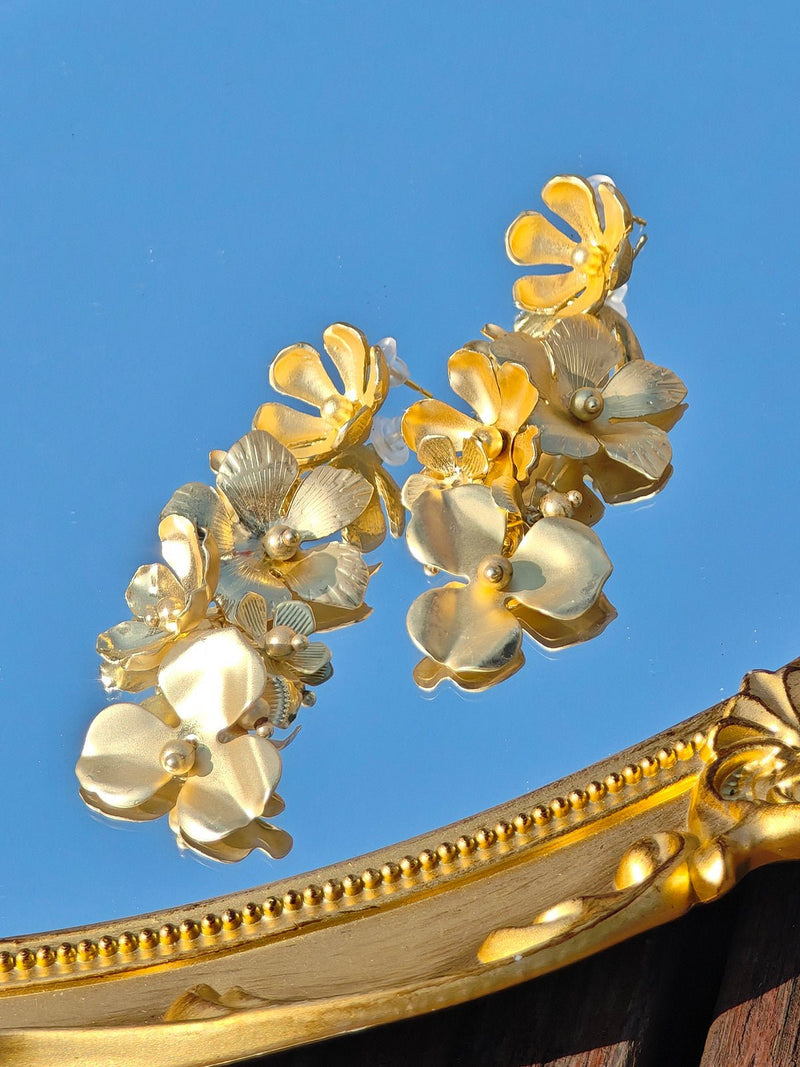 Social Queen's Flower Tassel Earrings, featuring a layered floral design with pearl accents and gold plating, are displayed on a reflective surface beside an ornate gold frame.