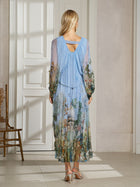 A woman faces away indoors, wearing the Social Queen Garden-Inspired Print Pleated Dress with long sleeves and strappy sandals. The ankle-length pleated dress features a nature print. A wooden chair sits on the left.