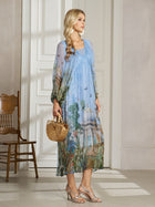 A woman stands indoors wearing the Social Queen Garden-Inspired Print Pleated Dress, paired with a wooden handbag, strappy heels, and long earrings. A wooden chair is visible in the background.