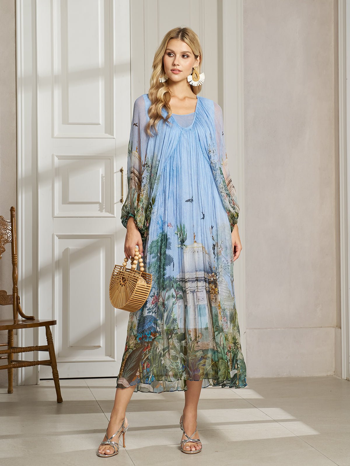 A woman stands indoors wearing the Social Queen Garden-Inspired Print Pleated Dress with long sleeves and a scenic design, accessorized with a small wicker bag, sandals, and large earrings.